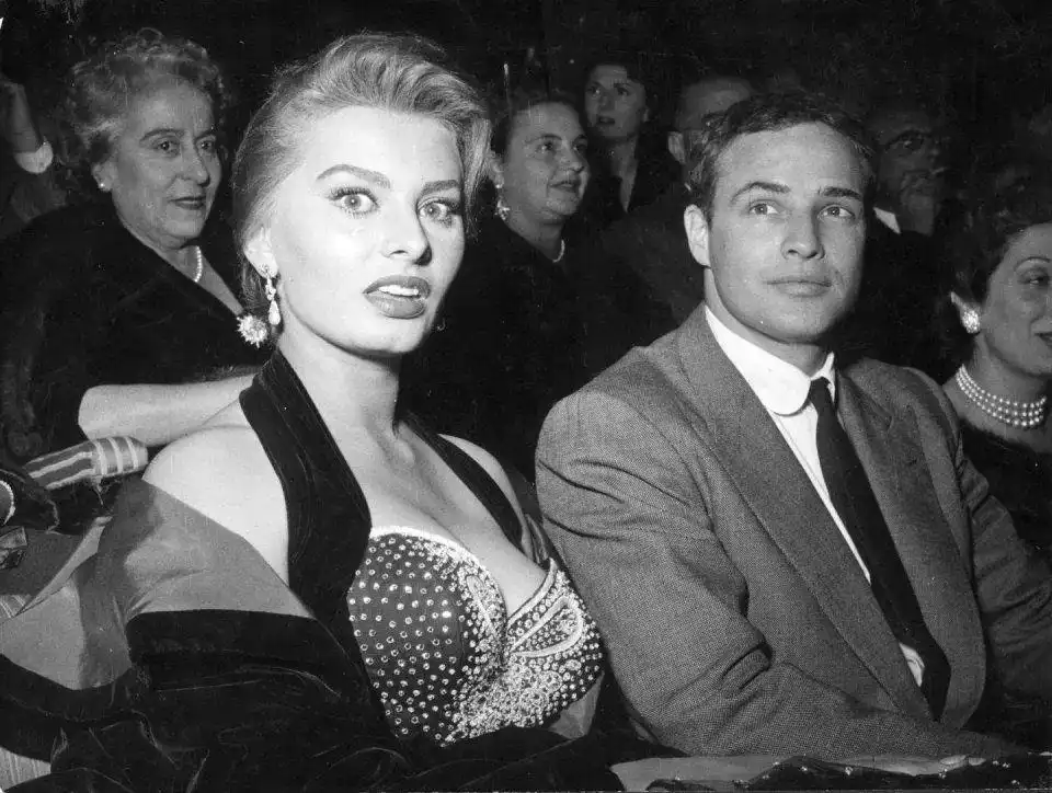 A black-and-white photo shows a glamorous woman in a beaded dress and a man in a suit seated next to each other in an…