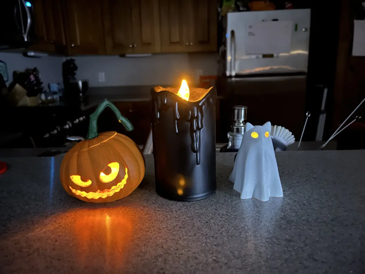 A dimly lit kitchen counter displays three Halloween decorations: a glowing jack-o'-lantern with a menacing grin, a black…