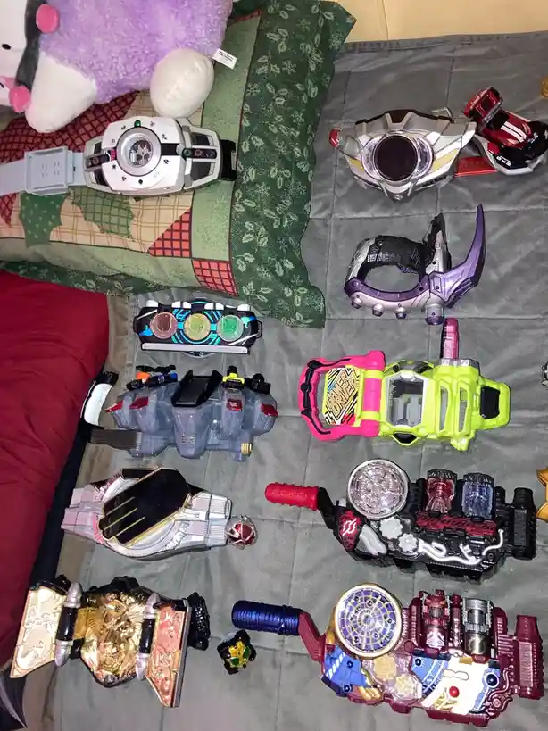An overhead shot displays a large collection of colorful, detailed Kamen Rider transformation belts and devices arranged on…