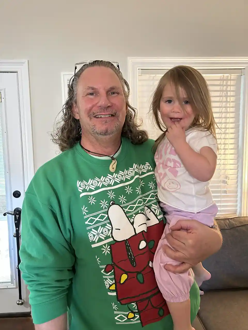 A man with long hair and glasses on his head wears a green holiday sweater with Snoopy and holds a young girl in pink pants…