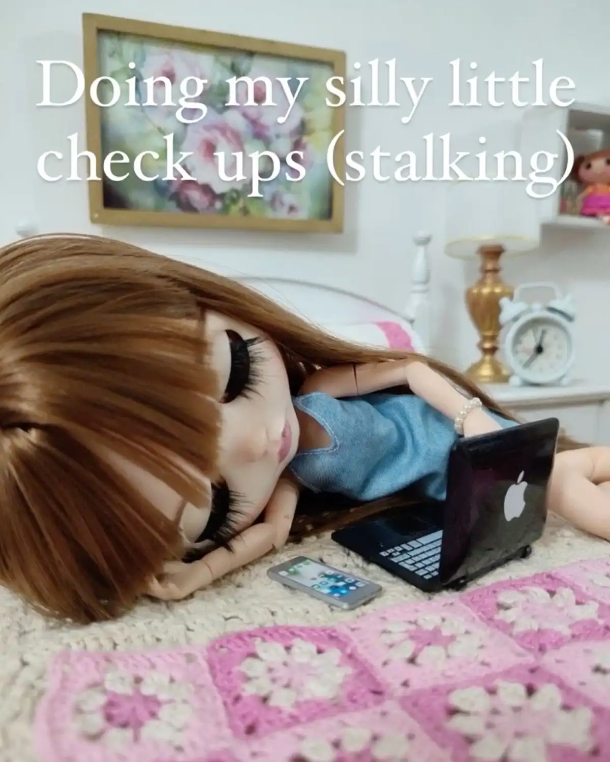 A doll with long brown hair lies on a bed, propped up on her elbow, looking at a miniature laptop. A tiny smartphone rests…