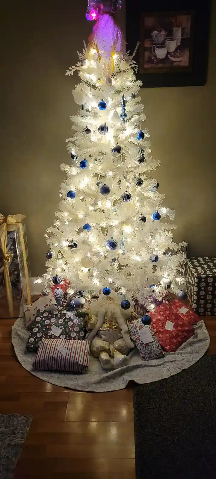 A full, decorated white artificial Christmas tree with blue ornaments and warm lights stands in a room, surrounded by…