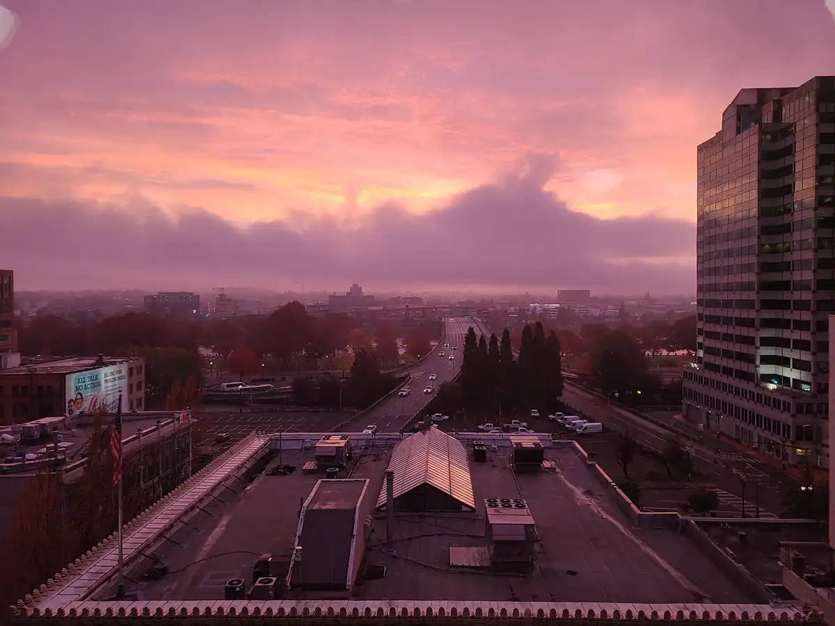 A cityscape at dawn or dusk with a vibrant pink and orange sky, featuring a highway with light traffic, buildings, and…