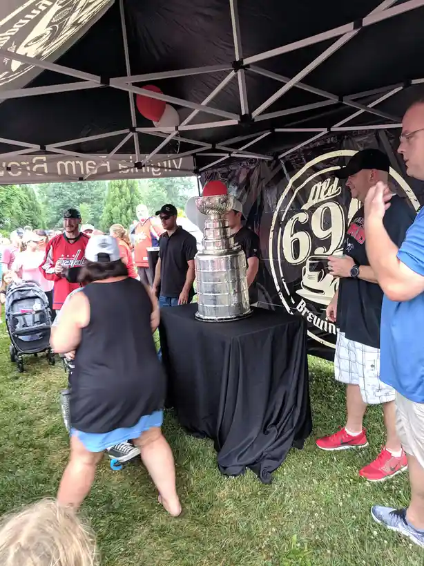 A large silver Stanley Cup trophy sits on a black-clothed table under a black pop-up tent at an outdoor event. Several…