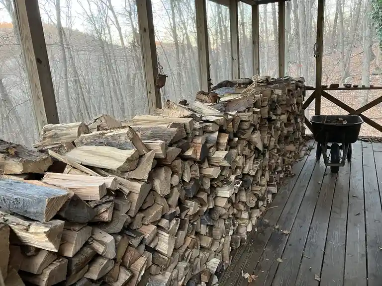 A large, neatly stacked pile of chopped firewood fills a wooden screened porch, with a black wheelbarrow nearby and a forest…