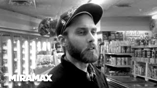 A black-and-white image of a bearded man in a baseball cap standing in a grocery store aisle, looking slightly to his right…