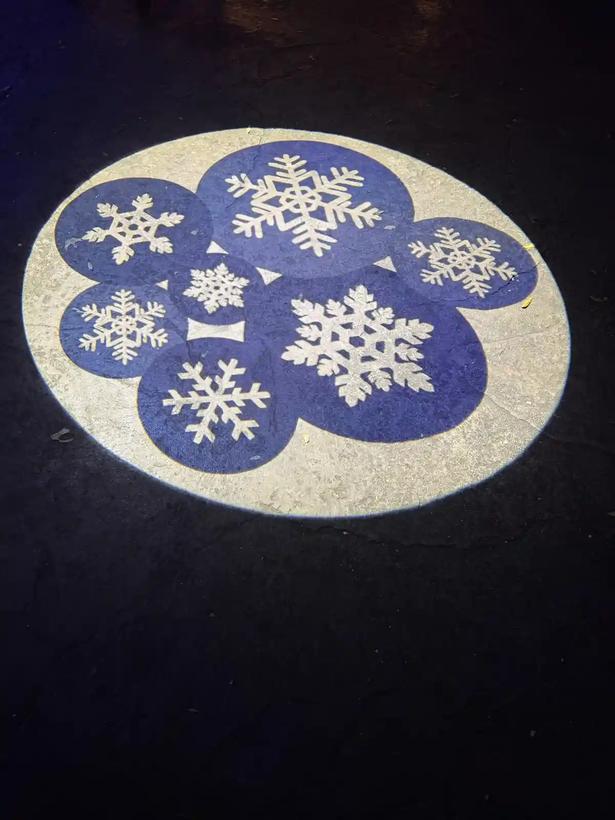 A circular pavement decoration with a light gray border and a dark blue center, featuring six white snowflake designs…