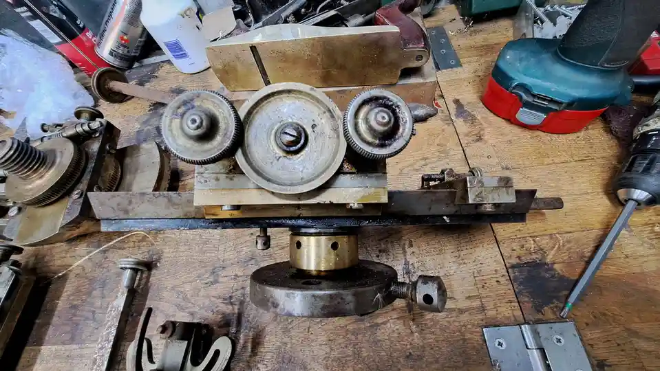 A cluttered workshop table holds a complex, homemade-looking machine with three interlocking gears and brass components…