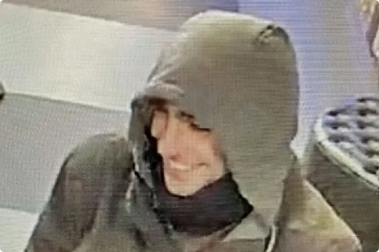 A low-resolution security camera image shows a person wearing a dark hooded jacket with the hood up, obscuring most of their…