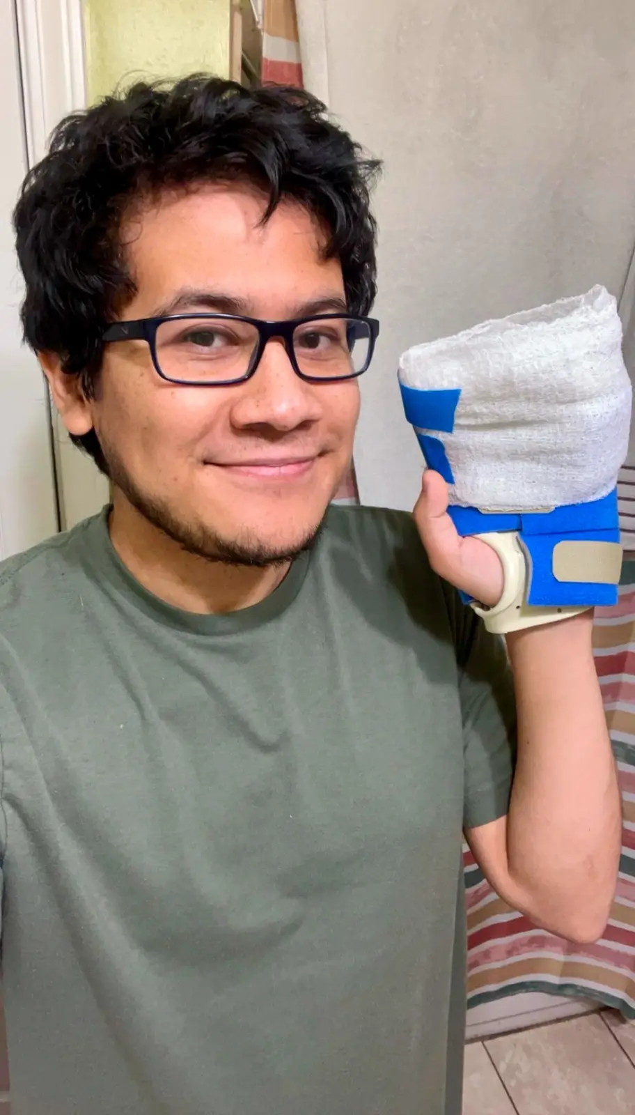A man with dark curly hair and black-rimmed glasses smiles at the camera while holding up his right hand, which is heavily…