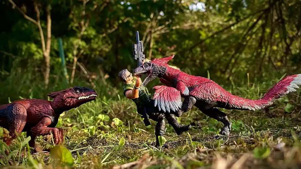 A toy action figure of a man holding a rifle is being attacked by a large red and black feathered dinosaur toy, while…