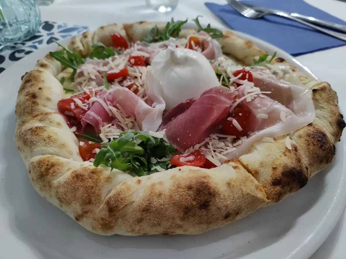A close-up photo of a round, flatbread-style dish on a white plate, topped with arugula, chopped tomatoes, thin slices of…