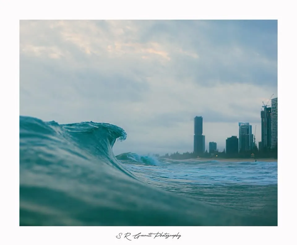 A low-angle photo of a large ocean wave curling toward the viewer, with a city skyline of tall buildings visible in the hazy…