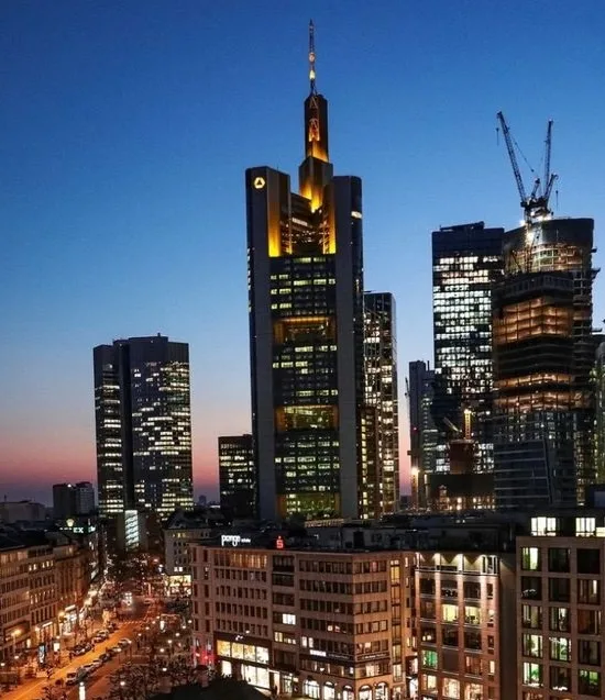 a dusk photograph of the frankfurt skyline, prominently featuring the illuminated kommerzbank tower with its distinctive…