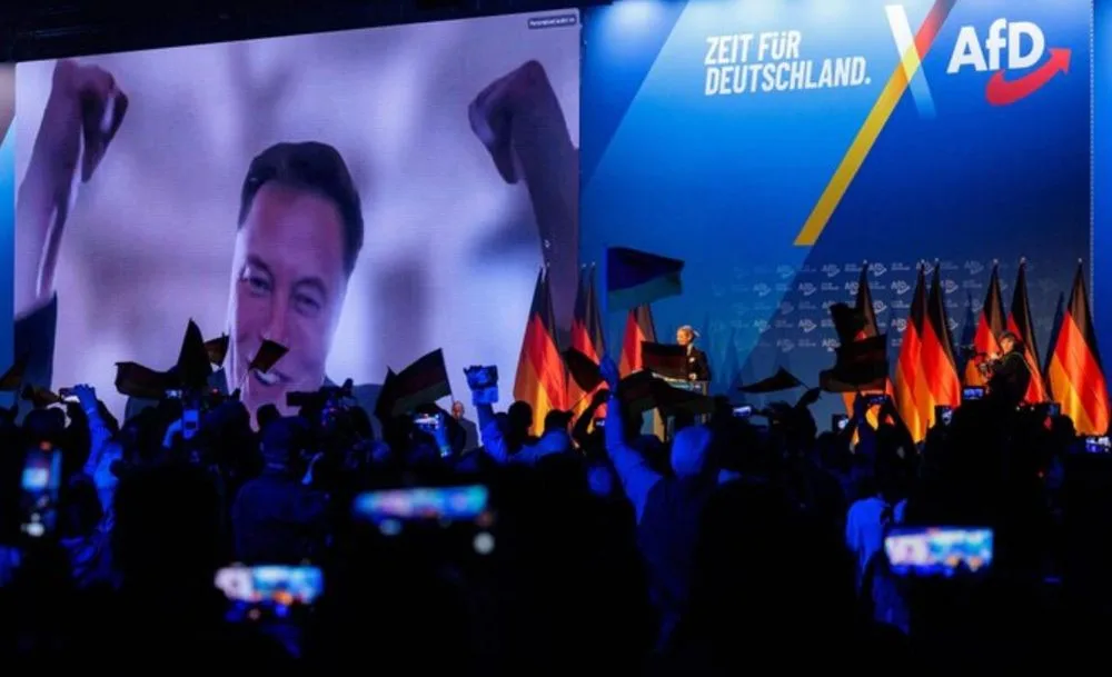 A large screen at an AfD (Alternative für Deutschland) political rally displays a smiling Elon Musk with raised fists, while…