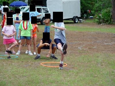 A group of children are playing a game of hula hoop relay on a grassy field. One boy in a grey t-shirt and dark shorts is…