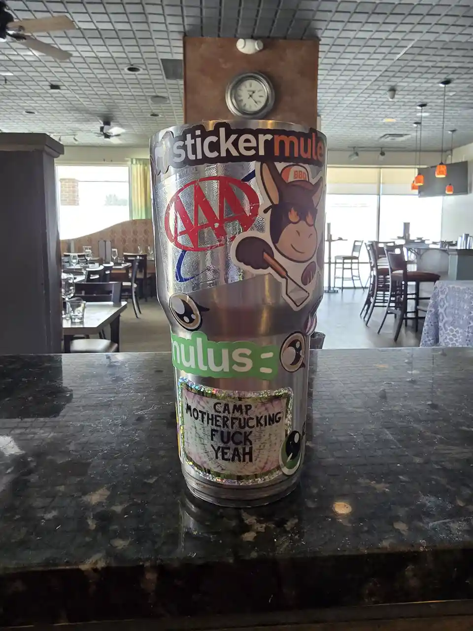 A heavily stickered silver tumbler sits on a dark countertop in a restaurant, covered with various decals including a…