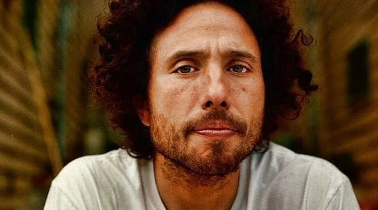 A close-up photo of a man with curly dark hair and a beard, wearing a white t-shirt, looking directly at the camera with a…