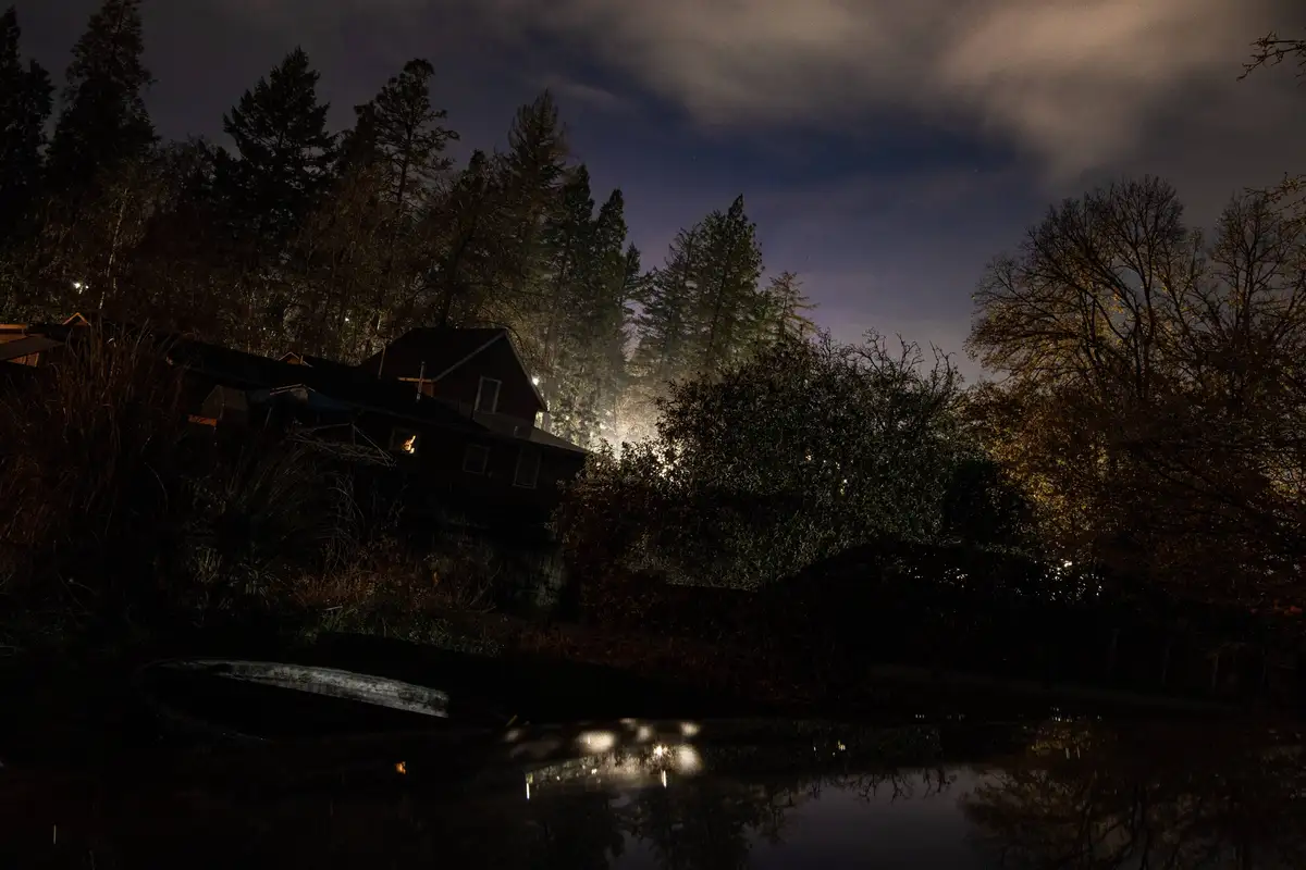 A dark, atmospheric night scene showing a house partially obscured by trees, with a body of water in the foreground…