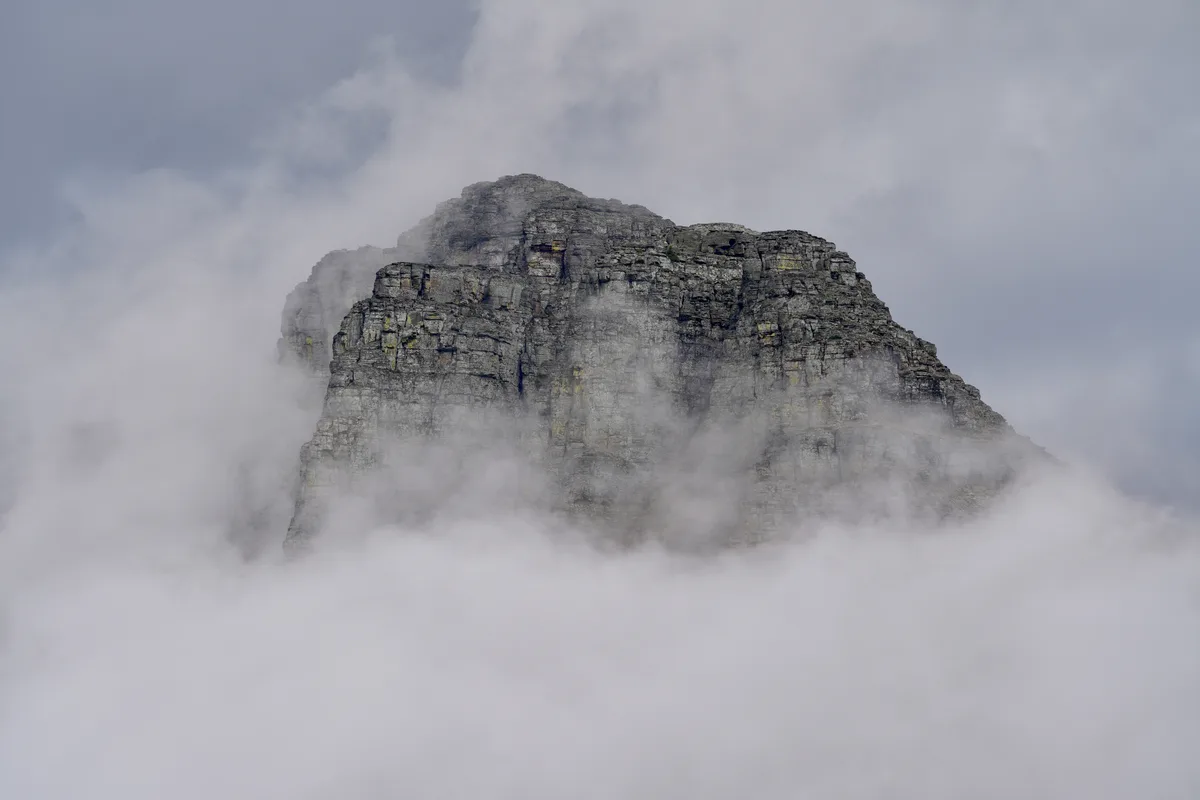 A rugged, rocky mountain peak emerges from a thick layer of clouds, with mist swirling around its base and partially…