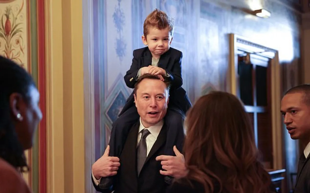 A man in a suit has a small child sitting on his shoulders, looking down with a serious expression. The child is wearing a…
