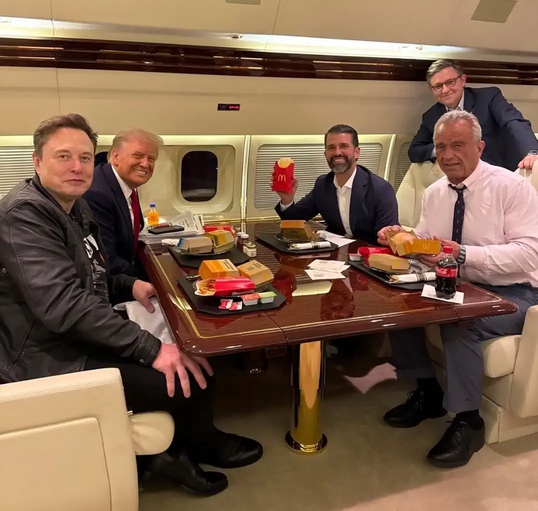 Five men, including Elon Musk and Donald Trump, are seated around a polished wooden table inside a luxurious private jet…