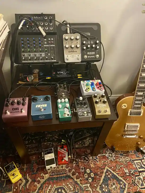 A cluttered wooden table holds a complex array of guitar effects pedals and a mixer, all interconnected with numerous…