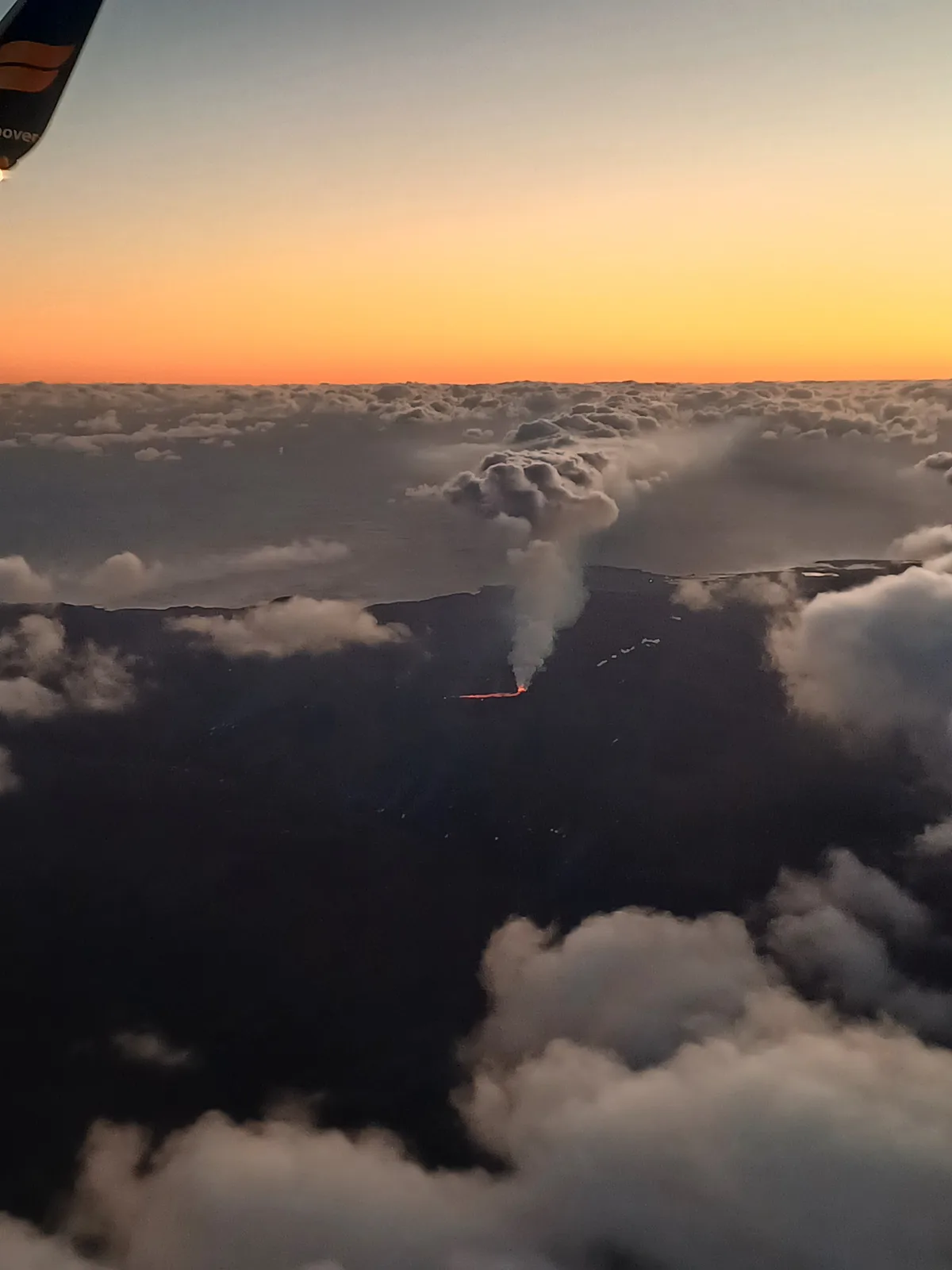 An aerial view from an airplane window shows a volcanic eruption below, with glowing orange lava flowing across a dark…