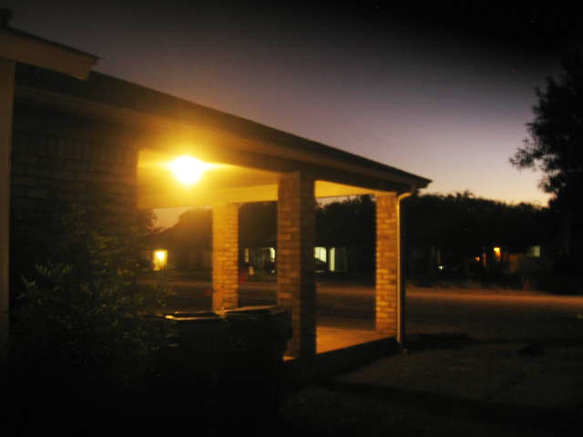 A dimly lit residential porch at dusk with a single bright overhead light casting a warm glow on brick pillars and a dark…