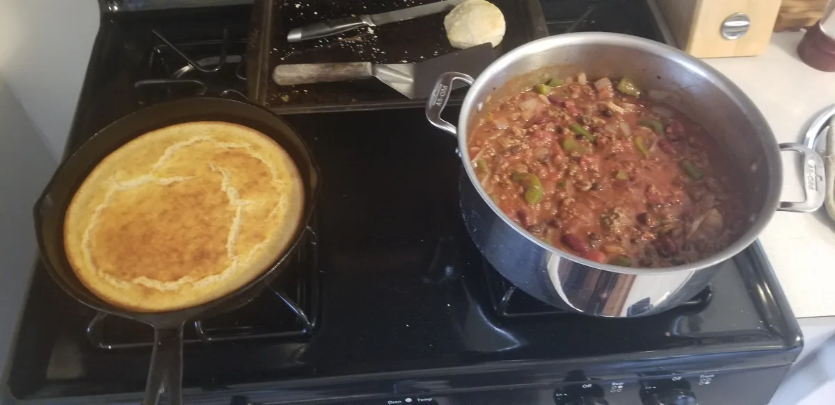 A kitchen stove with a cast-iron skillet holding a golden-brown cornbread on the left burner and a large pot of chili…