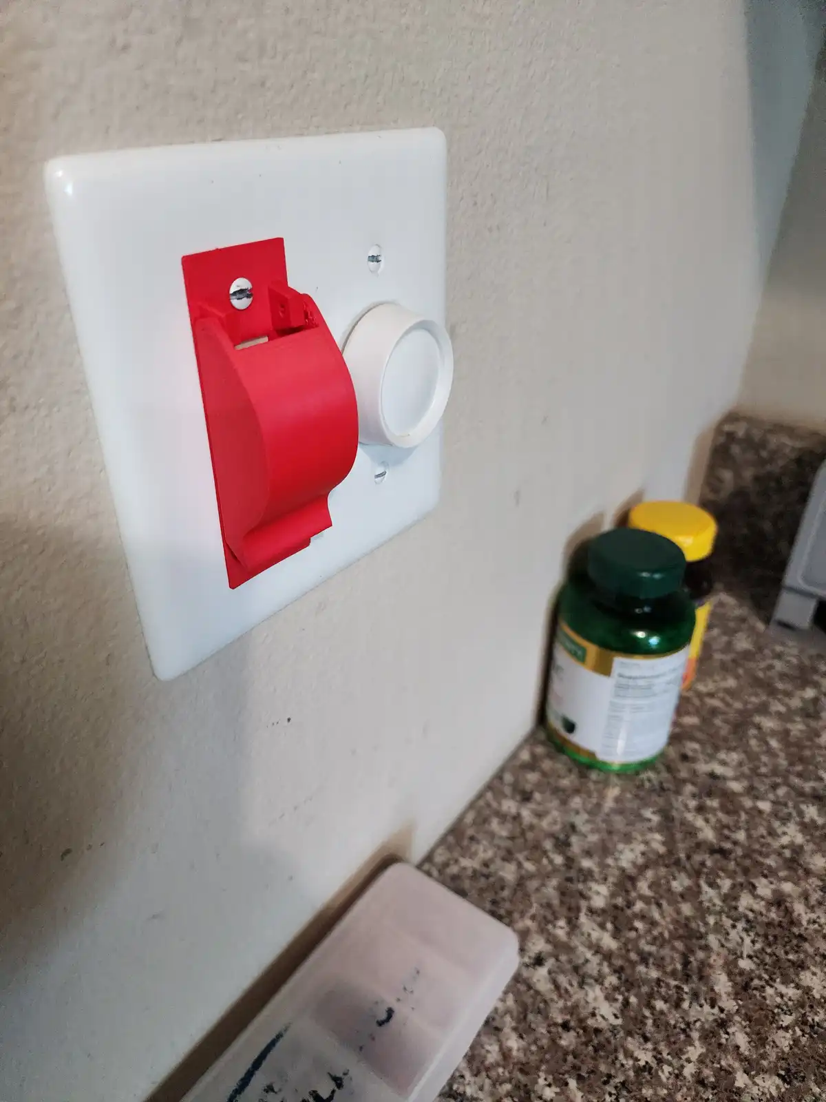A close-up photo shows a white wall-mounted light switch with a red plastic child safety cover partially covering it, next…