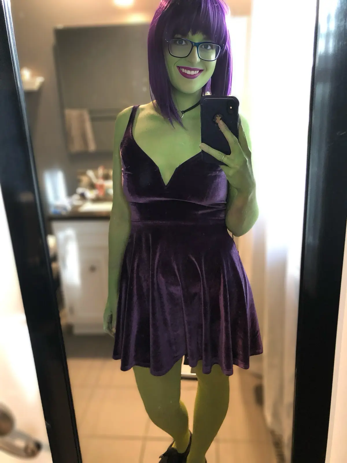 A person with green face paint, purple wig, and glasses takes a mirror selfie while wearing a purple velvet dress, holding a…