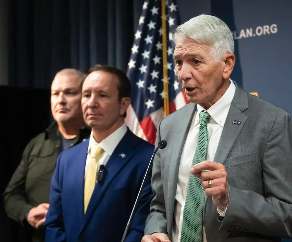 Three men stand at a press conference with an American flag behind them. The man in the foreground, wearing a gray suit and…