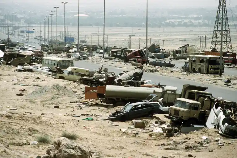 the image shows a wide, desolate road covered in sand and rubble, with dozens of wrecked vehicles including cars, buses…