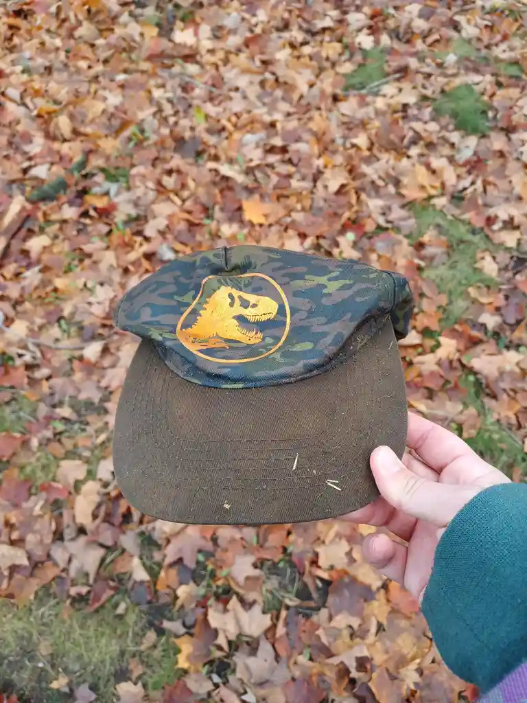 A hand holds a camouflage-patterned baseball cap with a yellow T-Rex skull logo against a backdrop of fallen autumn leaves…