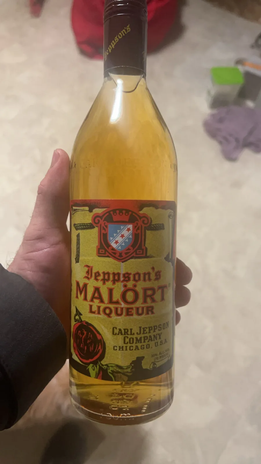 A hand holds a bottle of Jeppson's Malört liqueur, which has a distinctive yellow label featuring a crest, crossed swords…