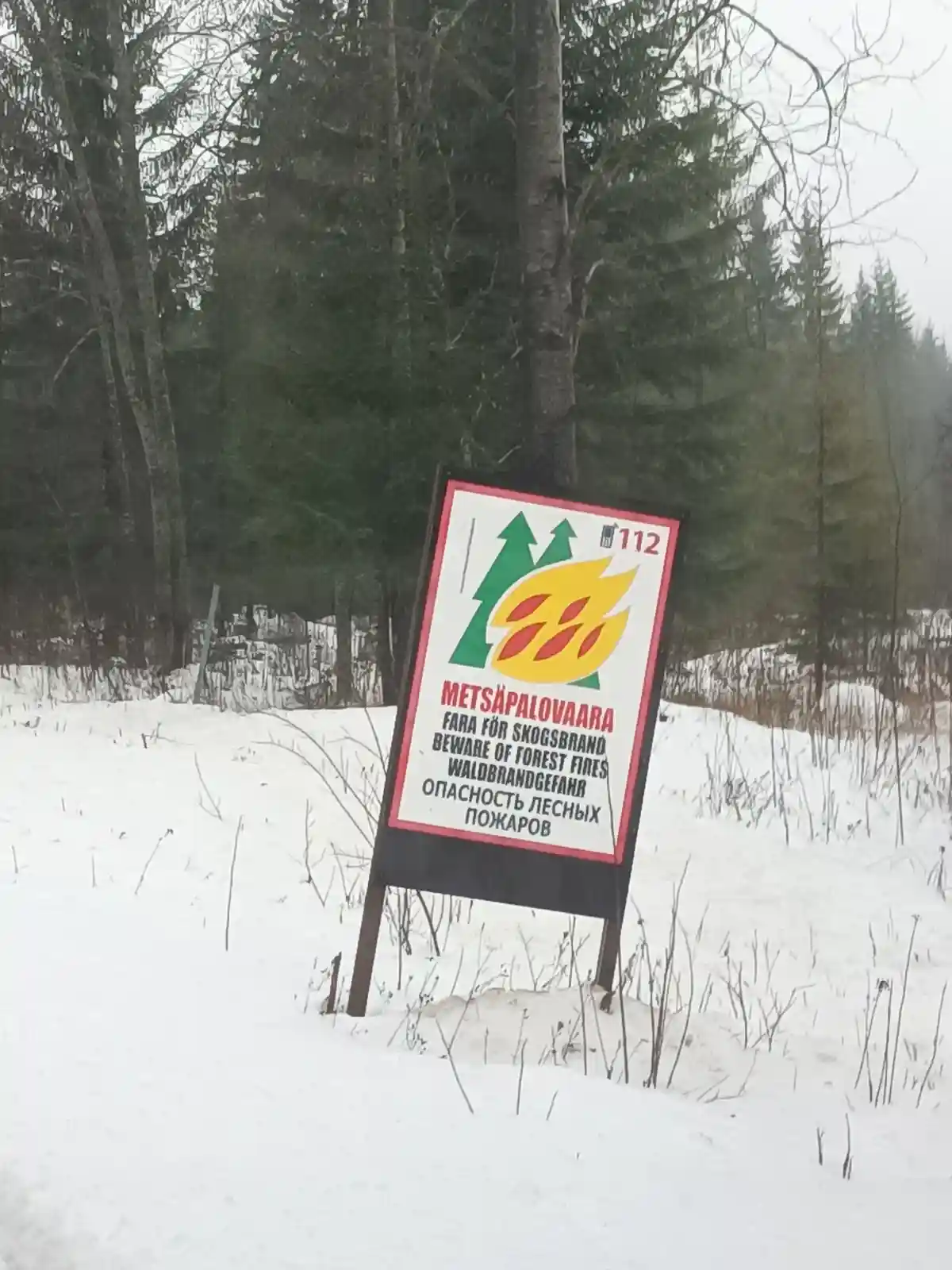 A multilingual forest fire warning sign stands in a snowy, wooded area, featuring green trees and a yellow flame graphic…