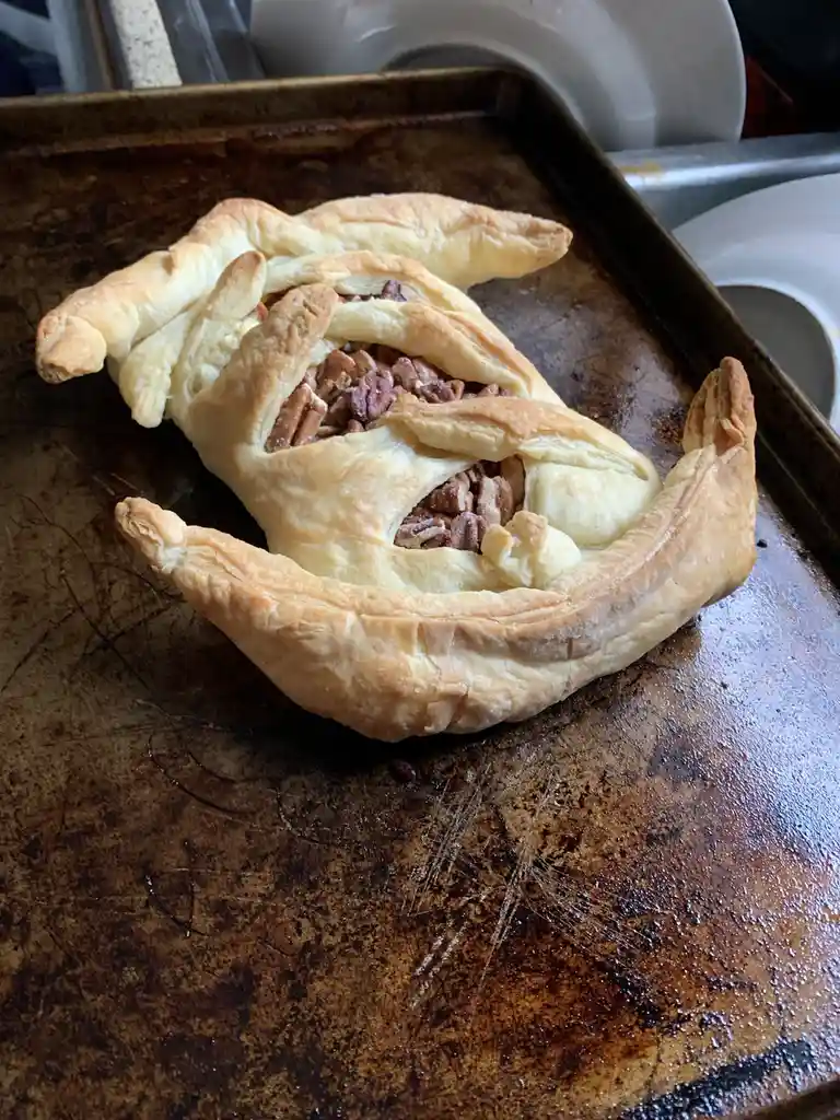 A golden-brown, baked pastry shaped like a dog, with pecan filling visible through the pastry's 'eyes' and 'mouth', sits on…