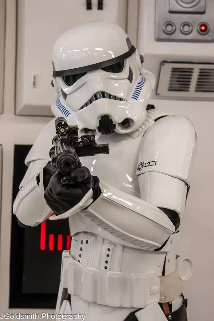A person in a white stormtrooper costume from Star Wars holds a black blaster pistol pointed forward. The image has text…