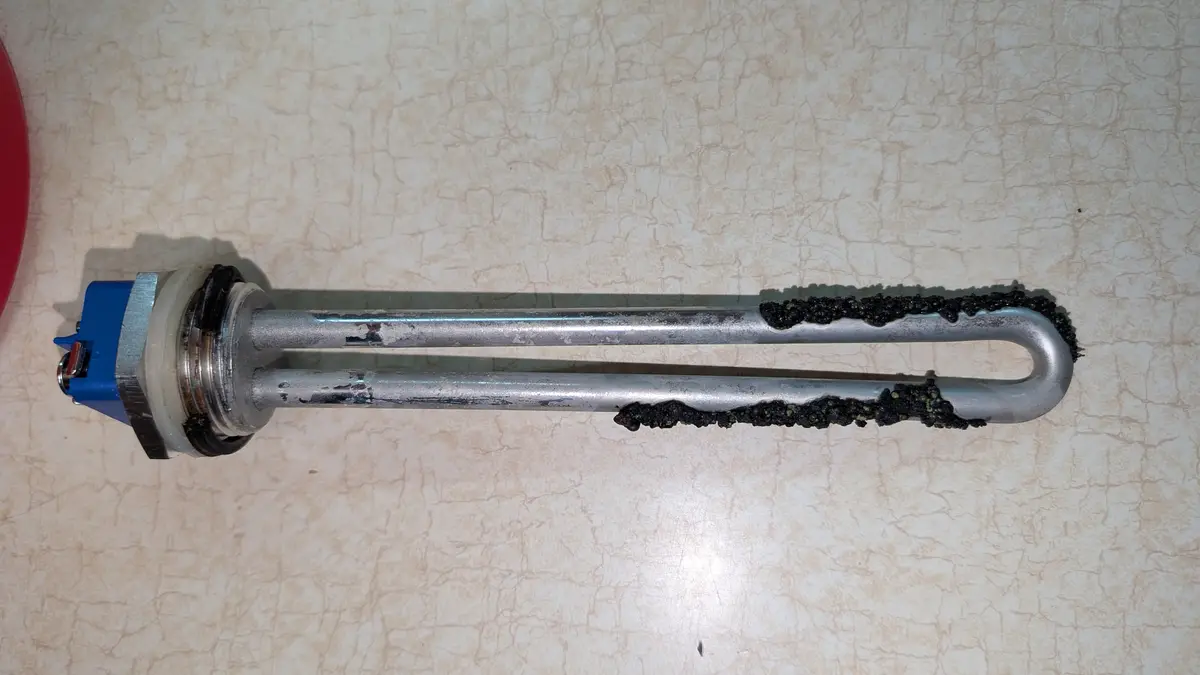 A close-up photo shows a metal immersion heater element lying on a beige, cracked-pattern countertop. The element has a blue…
