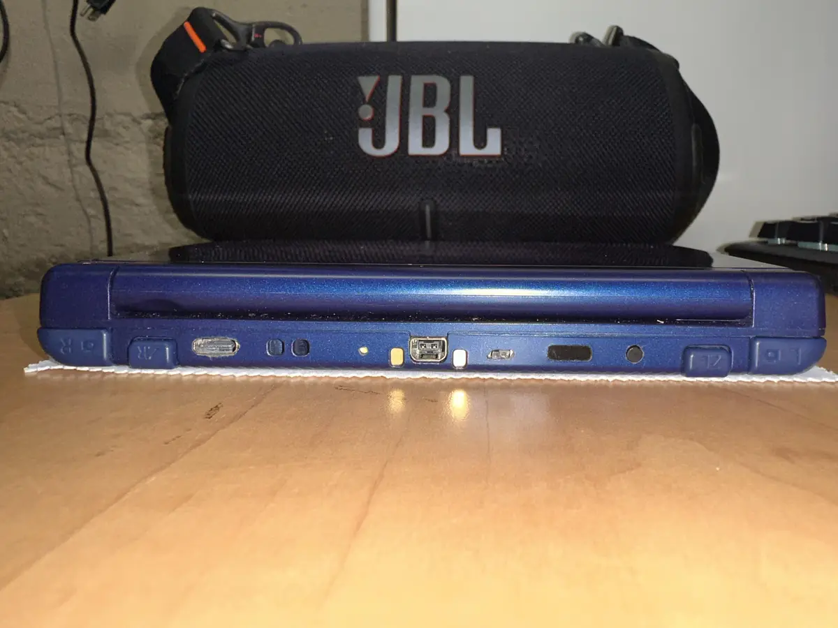 A close-up photo shows the side of a blue Nintendo DS Lite game console resting on a wooden surface, with its various ports…
