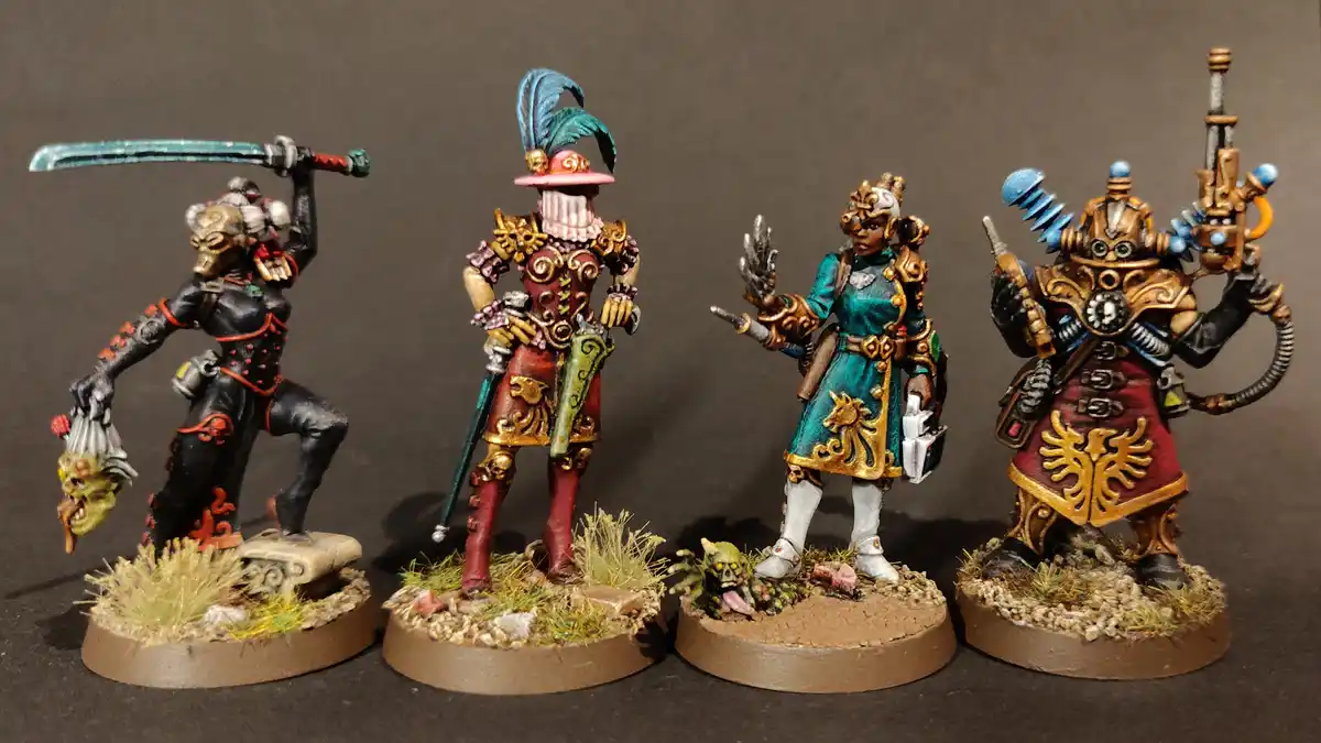 Four intricately painted fantasy miniatures stand side-by-side on textured bases against a dark background. From left to…