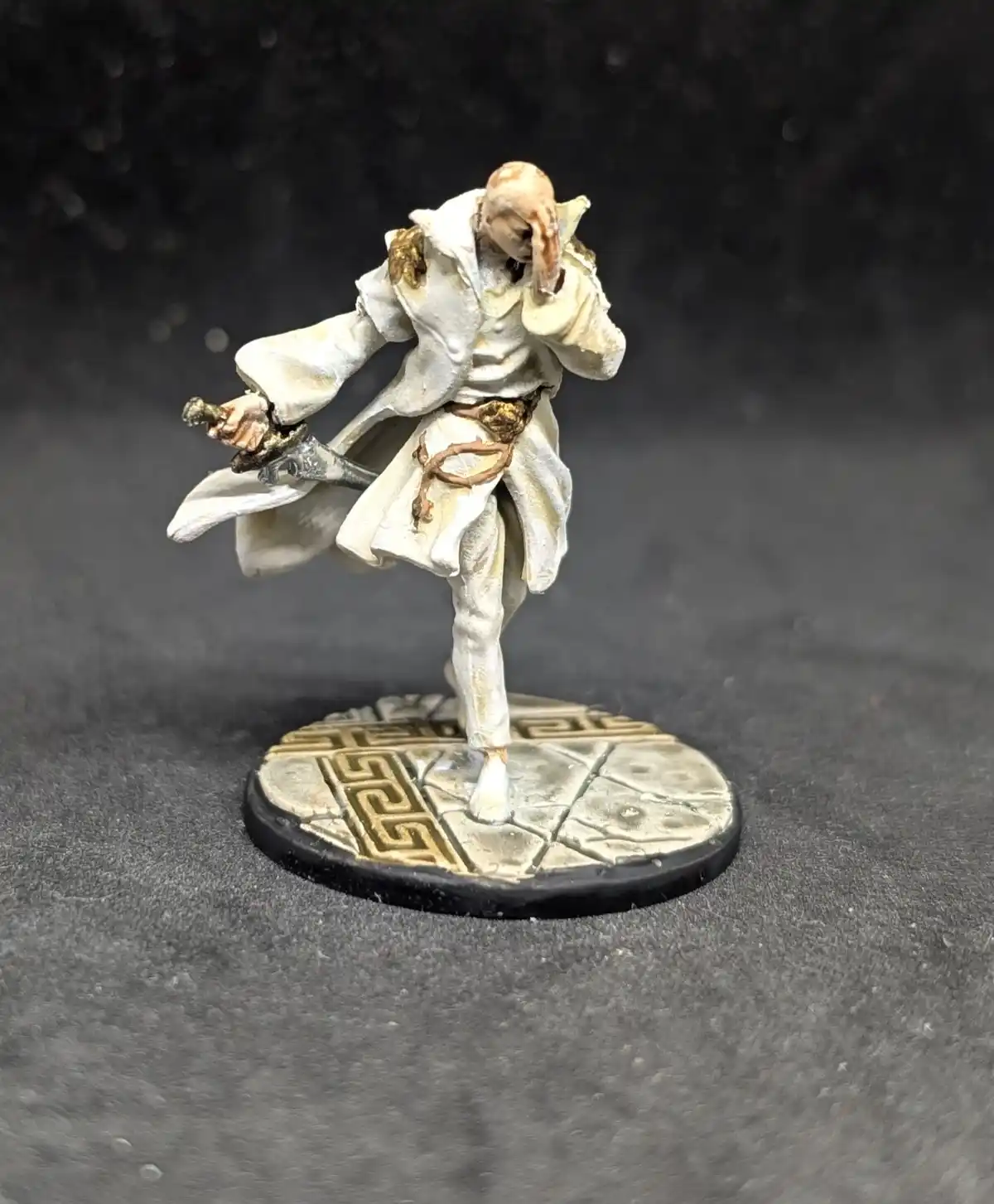 A painted miniature figure of a bald, white-robed warrior with a sword, standing on a patterned base, covers his face with…
