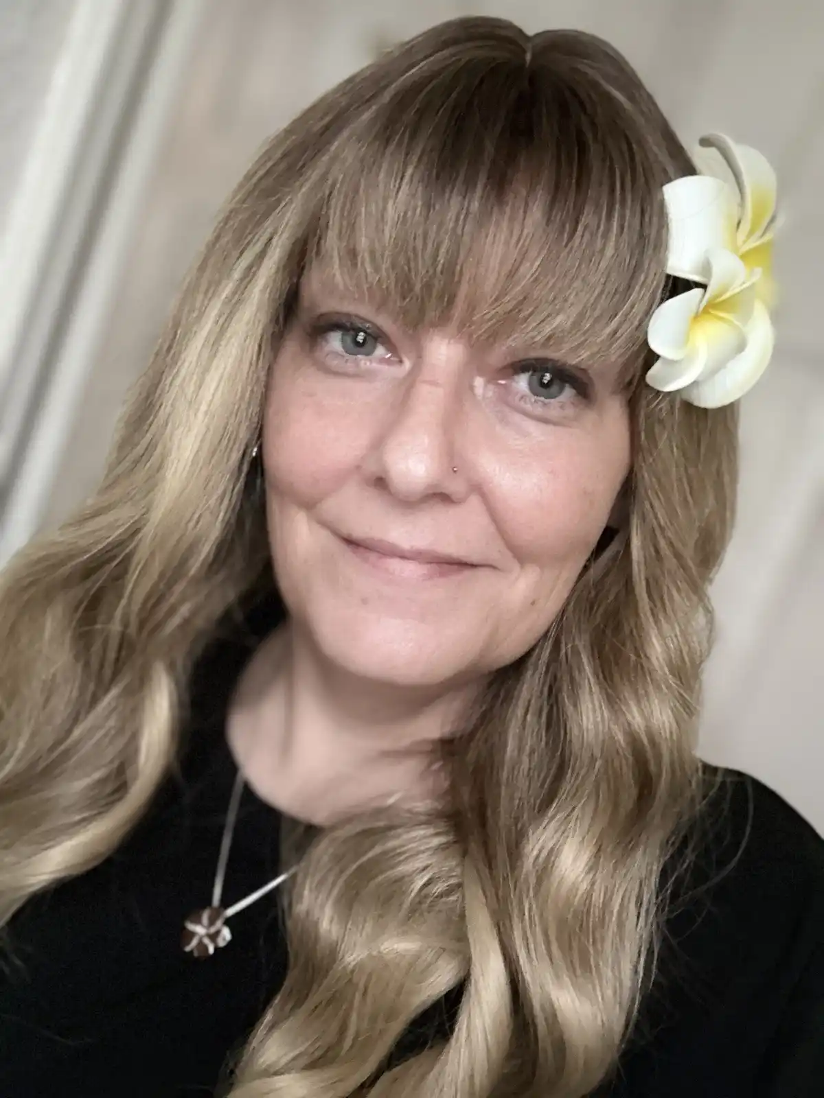A woman with long blonde hair and bangs smiles at the camera, wearing a white and yellow frangipani flower in her hair and a…