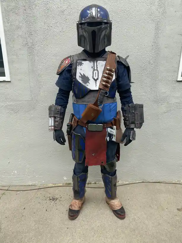 A person in a full, weathered Mandalorian armor costume stands facing forward against a plain gray wall. The armor is blue…