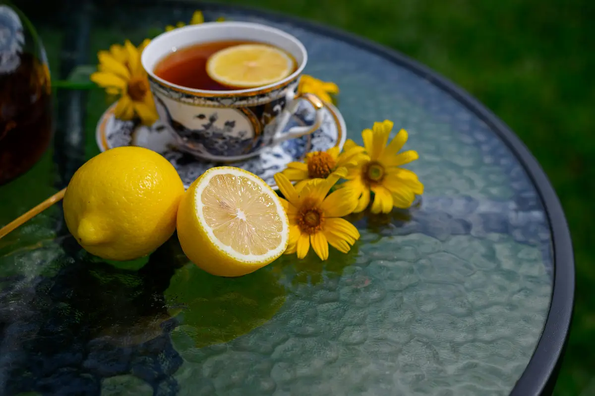 A serene outdoor scene features a decorative teacup filled with tea and a lemon slice, placed on a glass table alongside…