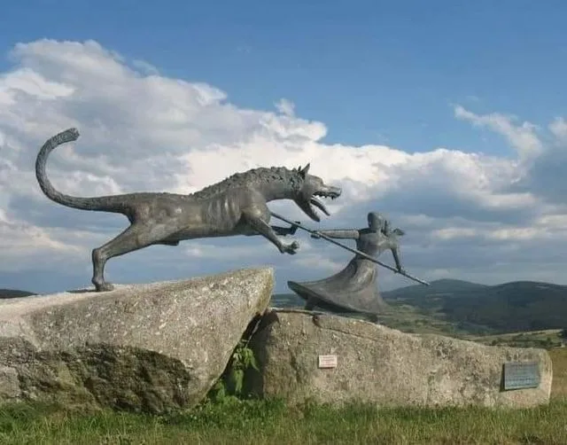 A bronze statue depicts a woman in a long dress holding a spear, standing on a large rock and confronting a large, snarling…