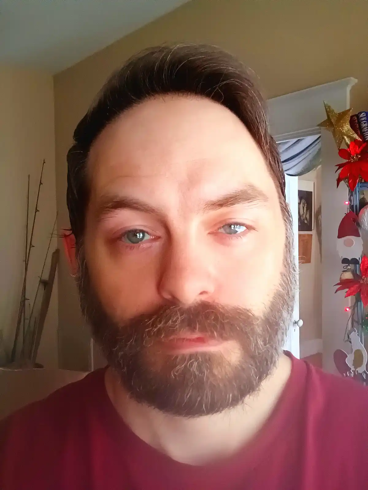 A man with a beard and blue eyes stares directly at the camera with a neutral expression, wearing a red shirt. Behind him, a…