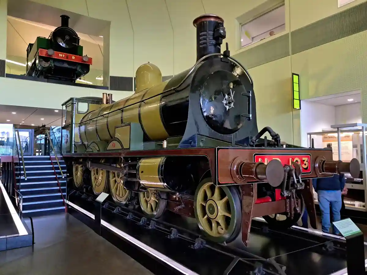 A large, vintage steam locomotive with green and gold paint is displayed indoors on a platform, with another smaller…