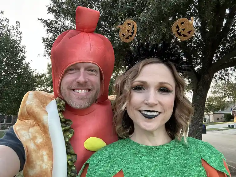 A man in a hot dog costume with a bun and mustard, standing next to a woman in a green pumpkin costume with black makeup and…